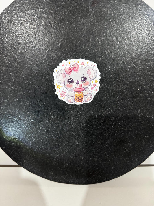 May include: A sticker featuring a cute cartoon koala with a pink bow, holding a boba tea. The koala has large, sparkling eyes and is surrounded by hearts and flowers. The sticker has a white border and is set against a dark background.