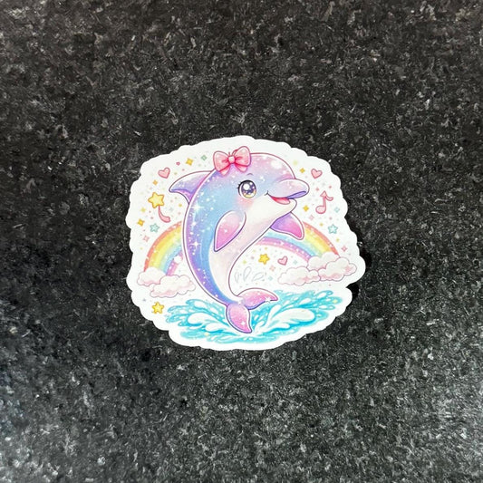 May include: A colorful sticker featuring a cartoon dolphin with a pink bow, swimming in blue water under a rainbow. The dolphin is primarily blue and pink, with stars and musical notes around the edges.